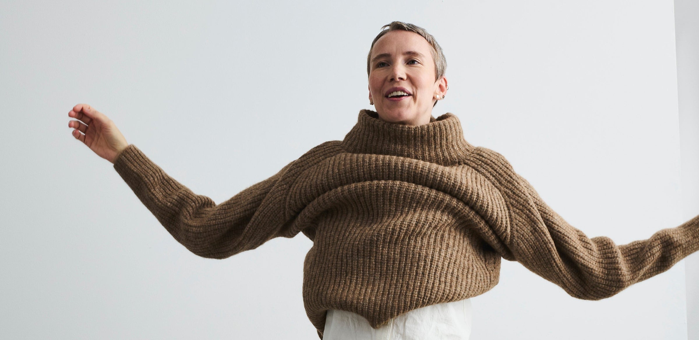 Person wearing a FOUR OBJECTS brown knitted sweater and beige pants against a white wall.