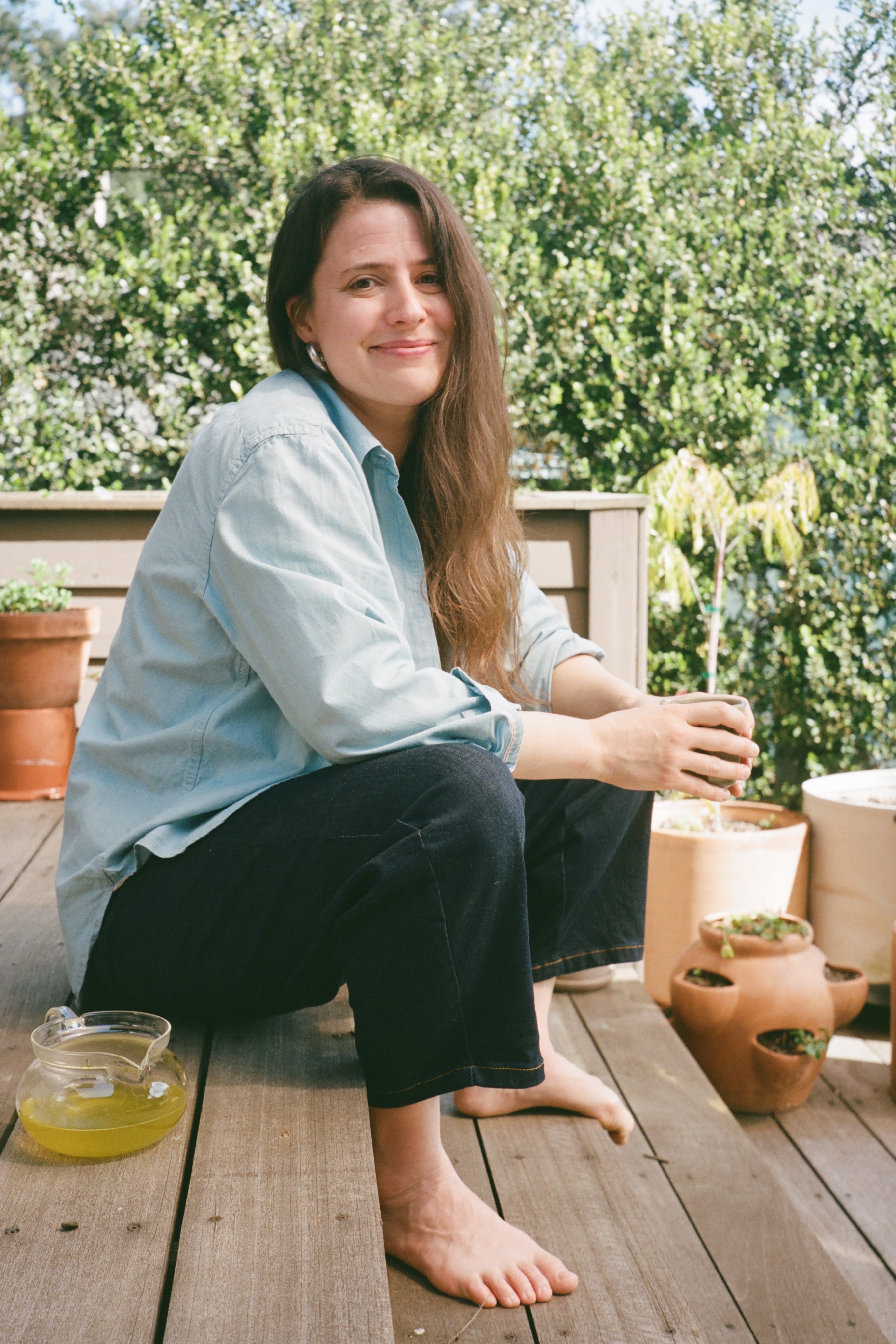 Fnny Singer wearing Four Objects chambray shirt sitting on a wooden deck with potted plants, holding a glass.