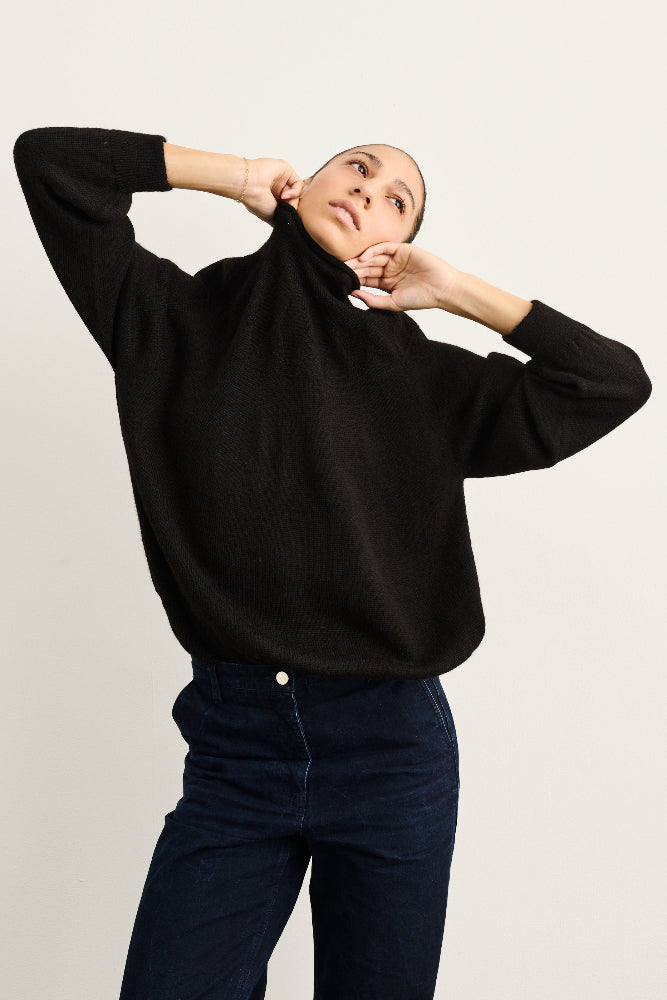 A woman wearing a black long-sleeve knit sweater and dark blue jeans, posing with her hands behind her neck against a plain light gray background.