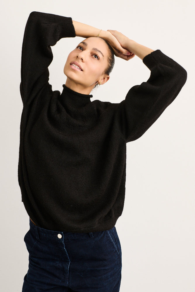A woman wearing a black long-sleeve knit mock neck sweater and dark blue jeans, posing with her arms raised against a plain light gray background.
