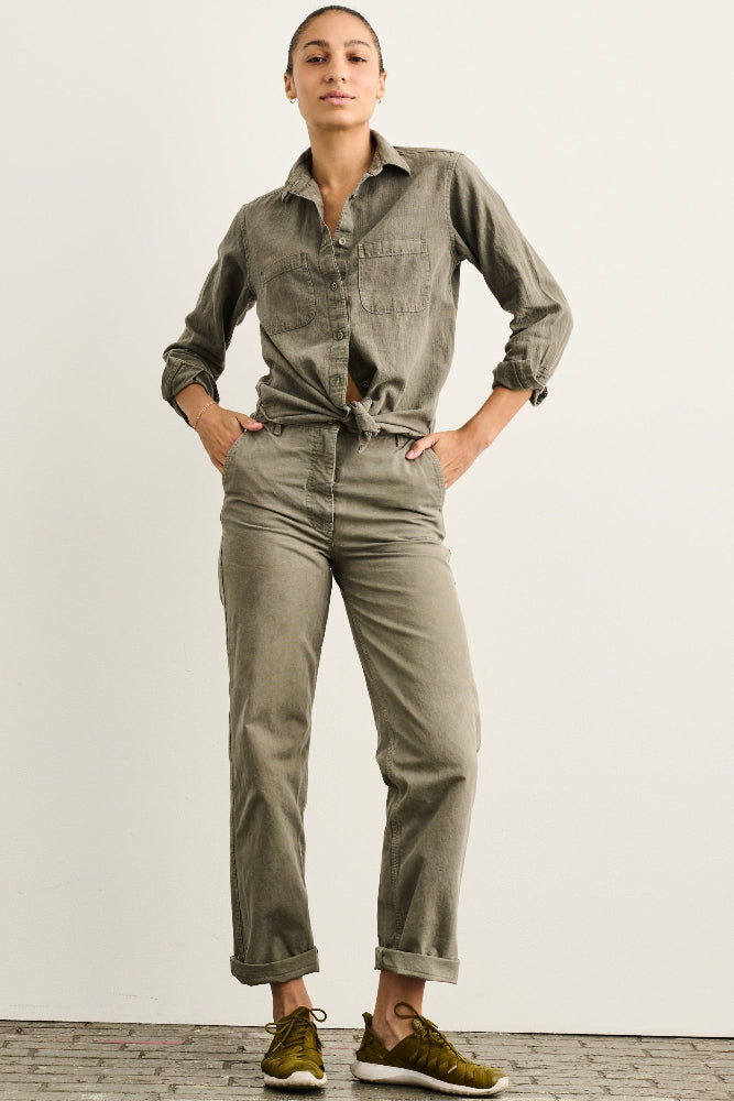 A model stands against a plain off-white wall wearing an olive green long-sleeve button-up shirt tied at the waist and matching olive green straight-leg jeans, paired with olive green sneakers.