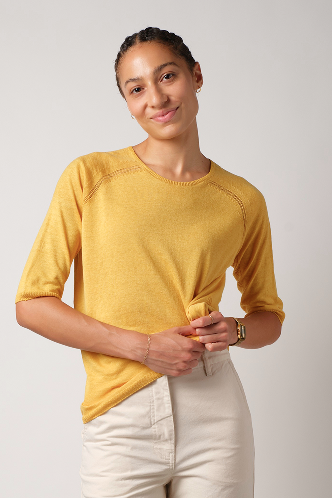 Woman wearing a yellow silk and linen short sleeve sweater and undyed organic cotton pants against a plain background