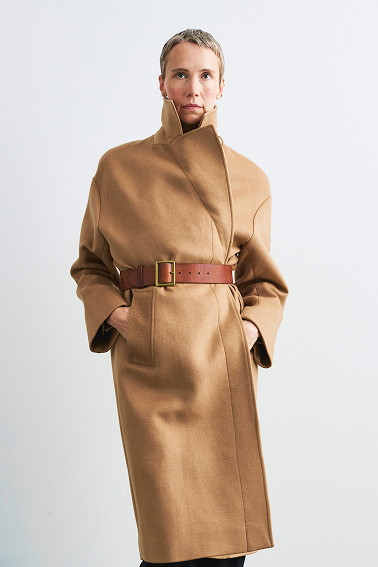 Person wearing a long camel coat bby four objects with a brown leather belt, standing against a white wall.