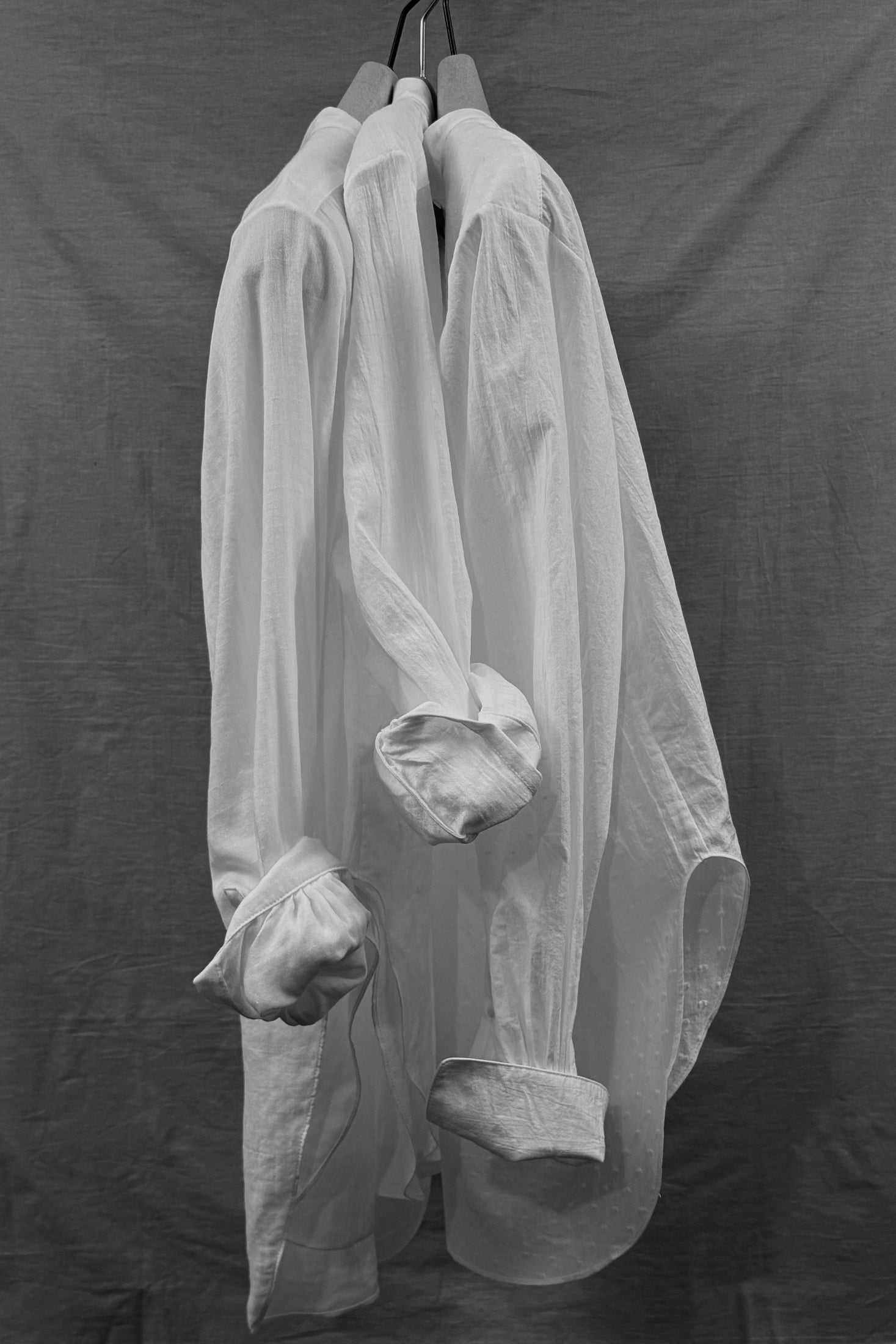 four objects organic cotton white shirts with sleeves rolled hanging against a dark background