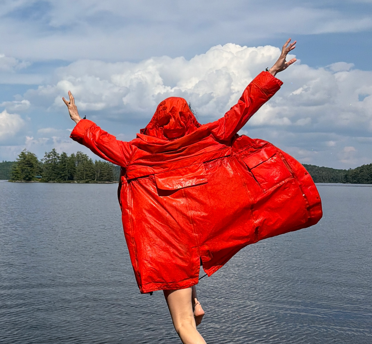 A person wearing a bright scarlet hooded organic cotton raincoat jumps off a wooden dock over a lake, with arms raised and the coat flying wide in the air against a cloudy sky.