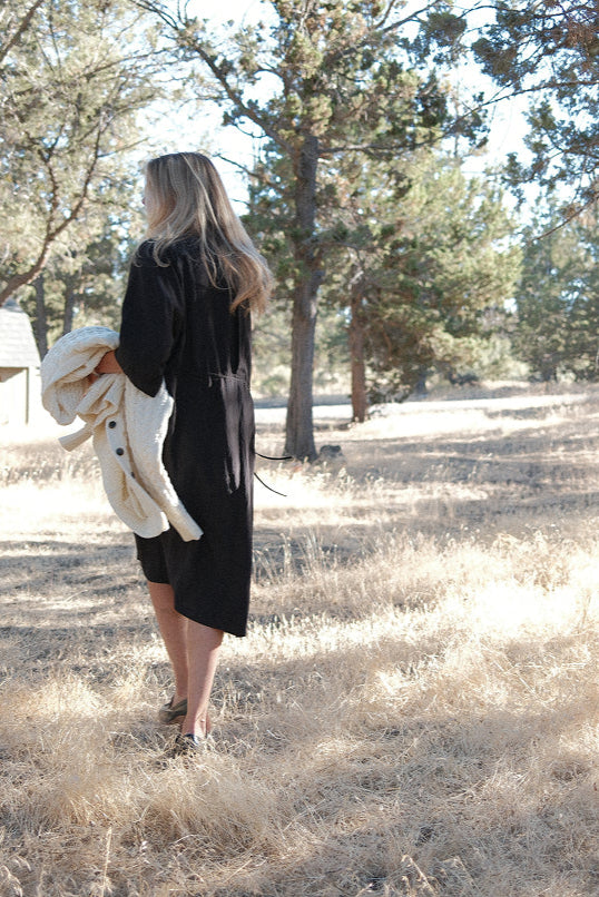 Person wearing a black silk FOUR OBJECTS dress holding an Aran Sweater by four objects in a forested area