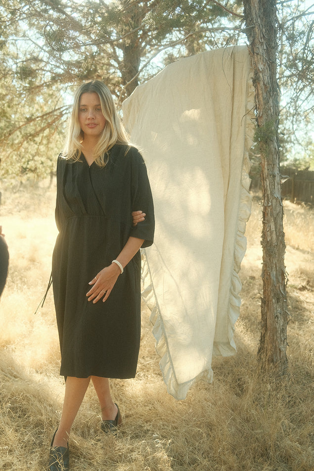 Woman in a FOUR OBJECTS SILK black dress standing outdoors with a white cloth draped over a tree