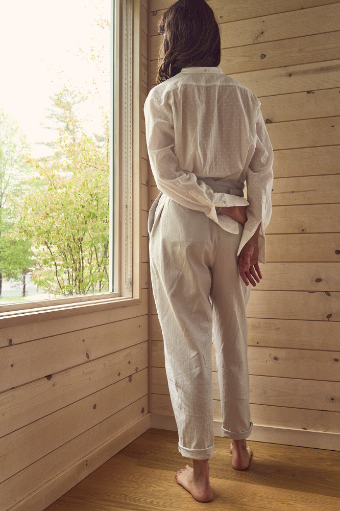 Person wearing a light-colored ORGANIC COTTON SHIRT OVER LINEN TROUSERS standing in a wooden room with a window.