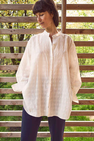 Woman wearing a white organic cotton windowpane blouse with a wooden lattice and greenery background