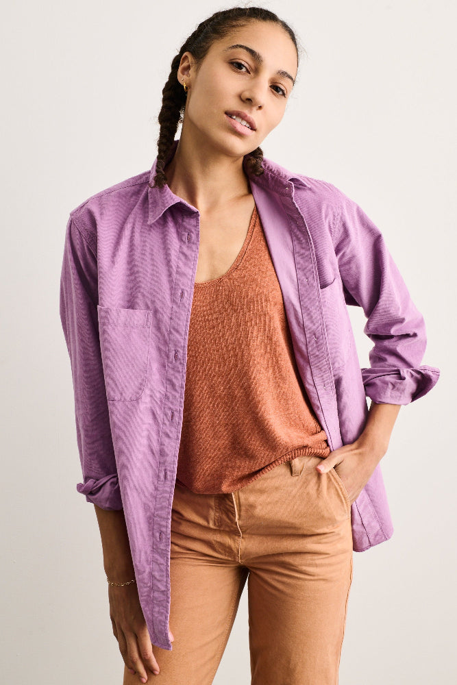A woman modeling a lavender-purple long-sleeve corduroy button-up shirt over a rust-orange tank top and tan pants, photographed against a plain light gray studio background.