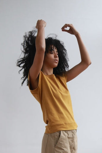 black women with big beautiful hair tossed up wearing a gold short sleeve tee over khaki chinos four objects organic cotton