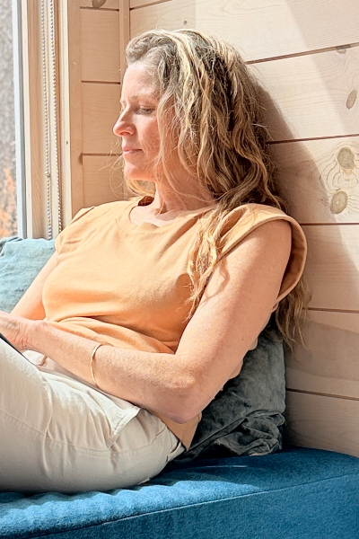 blond woman wearing an organic cotton tangerine tee sitting on a blue cushion against a wooden wall by a window holding a book.